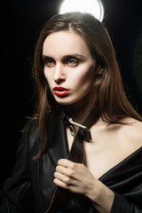 Braless girl with red lips, wearing a black unbuttoned blazer and a men's leather belt tightened around the neck, which she pulls with her hands,  on the dark background, next to a light lamp.