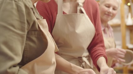 Tilting medium shot of young Caucasian female craft teacher in apron demonstrating hand sculpting technique for shaping clay bowl to elderly lady during pottery lesson