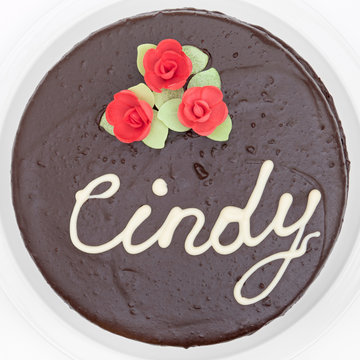 Birthday chocolate cake decorated with red roses for Cindy on white plate, top view