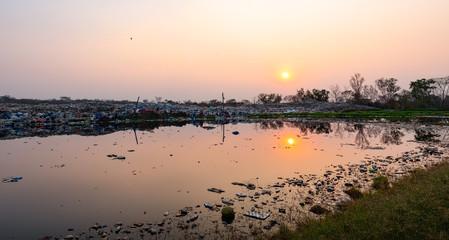 Polluted water and mountain large garbage pile and pollution at the sun is setting in the background