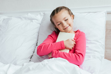 cheerful adorable kid hugging book while sitting on bed