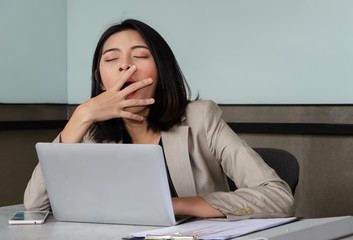 Young business woman yawning at meeting office table in front of laptop, covering her mouth out of courtesy. Overwork and sleep deprivation concept.