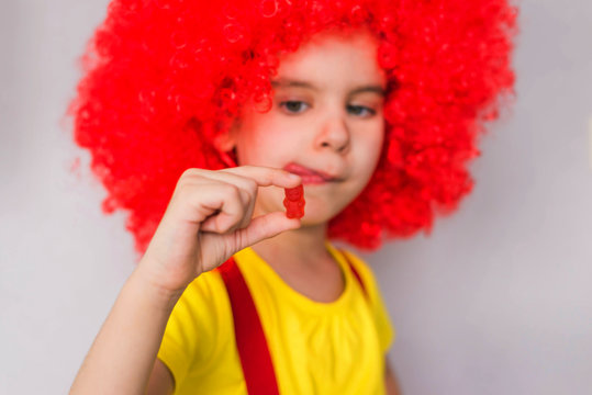 Little beautiful girl in a red wig holds a marmalade bear vitamin candy. Baby food, health concept.