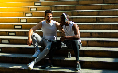  African american and friend white people sweating taking a break listening to music on phone after and  holding water bottle after running workout sitting on stair city