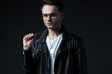 stylish brutal man in biker jacket holding pendant with token isolated on black