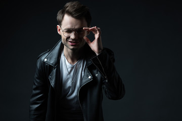 Obraz premium smiling stylish man in biker jacket touching eyeglasses and looking at camera isolated on black