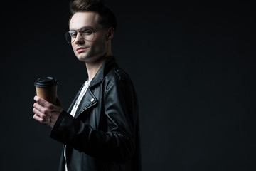 side view of stylish brutal man in biker jacket with coffee to go isolated on black