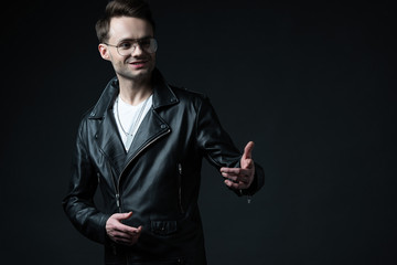 smiling stylish brutal man in biker jacket pointing with hand away isolated on black