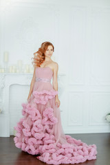 Female model in a lush pink wedding dress in full growth on a background of white room.