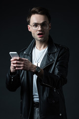 shocked stylish man in leather jacket using smartphone isolated on black