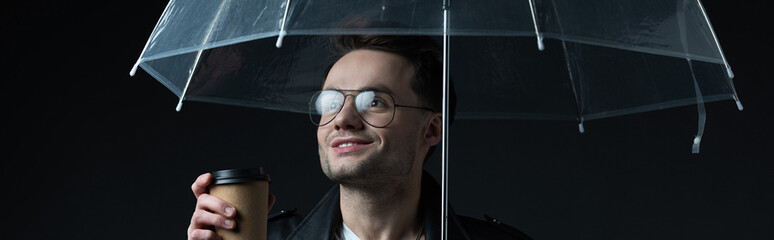 smiling stylish brutal man in biker jacket with umbrella and coffee to go isolated on black, panoramic shot