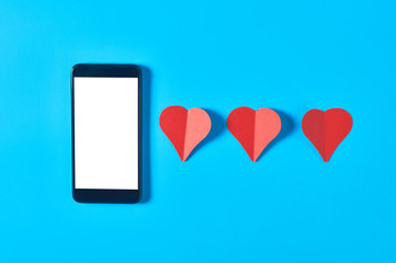 Black smartphone with isolated white screen for text, picture, photo and other graphics near row of three red paper hearts on blue table. Valentines day and love concept. Space for text. Top view