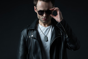 stylish brutal man posing in biker jacket and sunglasses isolated on black