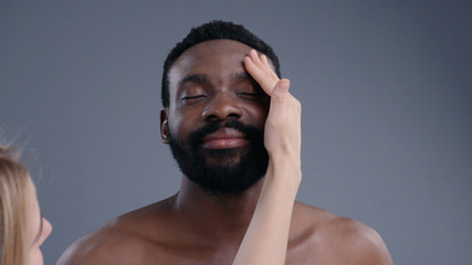Smiling portrait of satisfied african american nude young man on grey background. Make-up artist in process touching male model face preparing skin for shooting in the studio.