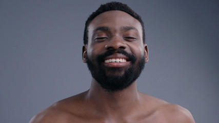 Portrait of happy attractive shirtless athletic male model applying perfume on fresh aftershower body enjoying aroma smiling at camera isolated on grey background.