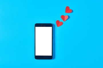 Black smartphone with isolated white screen for text, picture, photo and other graphics near three red paper hearts on blue table. Valentines day and love concept. Space for text. Top view