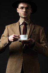 elegant man in beige suit holding coffee cup and saucer isolated on black