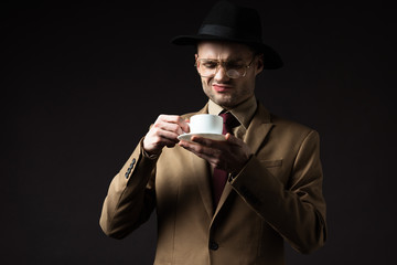 skeptical elegant man in beige suit, hat and eyeglasses holding coffee cup and saucer isolated on black
