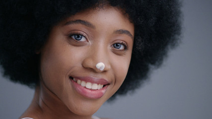 Close-up beauty portrait of a pretty african girl bare shoulders with moisturizing cream on her nose smiling on camera on grey background.