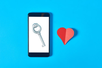 Black smartphone with isolated white screen for text, picture, photo and other graphics with vintage key near paper heart on blue table. Secrets or forbidden love concept. Top view