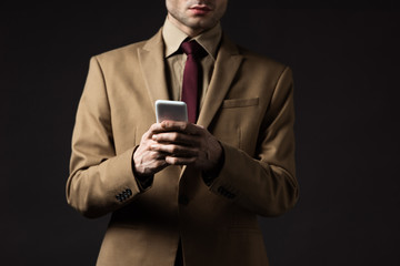cropped view of serious elegant man in beige suit using smartphone isolated on black