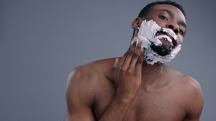 Close-up of handsome young afro-american shirtless man shaving and applying cream on his face....