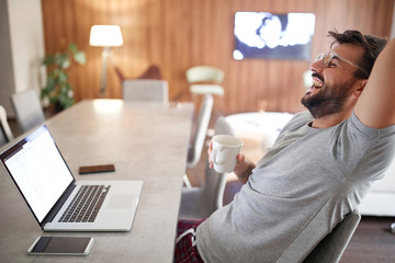 Smiling man drink morning cup of coffee while working on laptop.