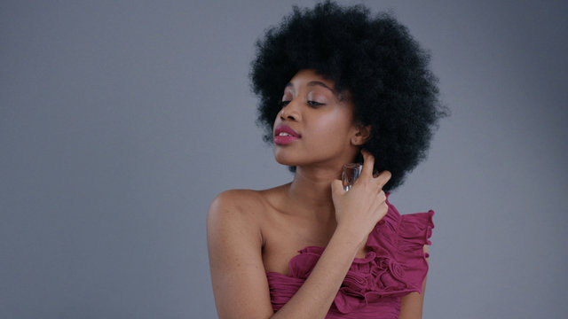 Amazing Black Young Woman With Afro Hairstyle Using A Lovely Women's Perfume On Grey Background. Portrait Of Sexy Female Black Model In Dress Trying A Fragrance Indoors.