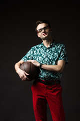 smiling fashionable man in eyeglasses in blue colorful shirt and red pants holding basketball isolated on black