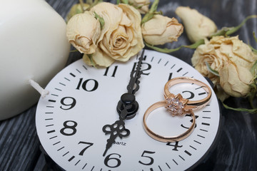 Wedding rings lie on the dial of the watch. Near a candle and dried roses. On brushed pine boards painted in black and white.