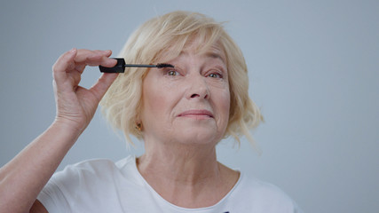 Close-up of gorgeous senior woman with blonde hair applying mascara on eyelashes. Pretty older woman doing make-up posing in the studio.