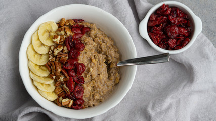 Chocolate oatmeal porridge with banana, pecan, nuts, dry cranberries in a white bowl on a grey napkin. There is a spoon in a bowl and little white bowl with some dry cranberries. 