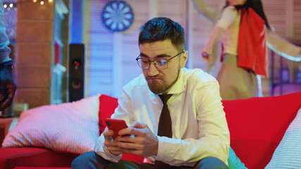 Handsome unsociable guy using social media or browsing news on smartphone getting upset on night party. People in background having fun celebration.