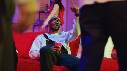 Fototapeta premium Drunk funny african american guy resting on couch tasting drinks sharing happy gestures with friends watching others dancing at cool night party.