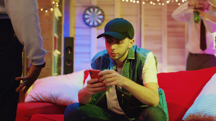 Lonely modern boy in hat browsing app on smartphone sitting on sofa looking around people dancing into music on background. Unsociable shy person.
