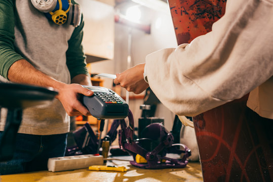 cropped view of worker holding terminal and woman paying with credit card and holding snowboard in repair shop