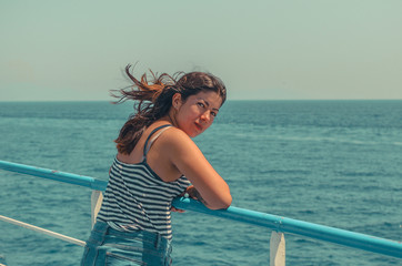cheerful young girl with black hair in a striped t-shirt and blue jeans stands on the deck of a ship at sea.