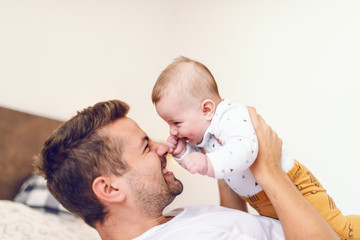 Handsome young dad lying in bed in bedroom and lifting his loving laughing baby boy six months old....