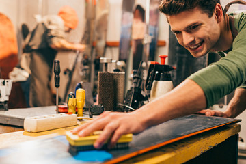 smiling worker using brush on snowboard in repair shop