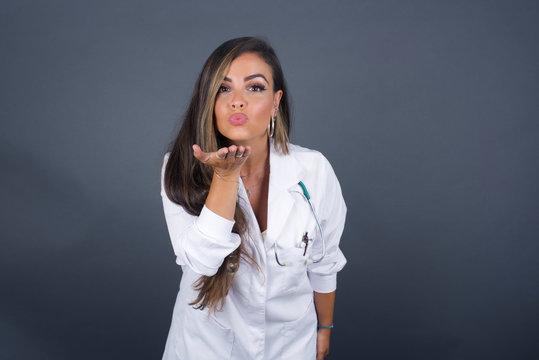 Cute appealing doctor female blows kiss at camera, demonstrates love to boyfriend or says goodbye on distance, isolated over gray studio background.