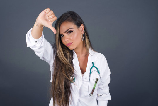 Discontent European doctor woman shows disapproval sign, keeps thumb down, expresses dislike, frowns face in discontent, isolated over gray background. Body language concept.