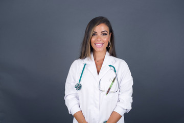 Beautiful Young  caucasian doctor woman with happy and funny face smiling and showing tongue. Wearing medical uniform and standing against gray studio background.