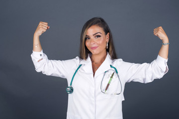 Waist up shot of caucasian doctor woman raises arms to show her muscles feels confident in victory, looks strong and independent, smiles positively at camera, stands against gray background.