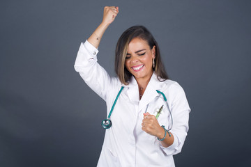 Attractive young caucasian doctor woman celebrating a victory punching the air with her fists and a beaming toothy smile over a gray studio background with copy space