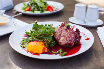 Grilled meat steak with cranberry sauce, glazed peaches and arugula salad on white plate.