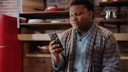 Serious sad young african american man use phone sitting at kitchen coffee tea serious...