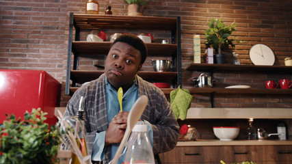 Funny handsome african american man with green spoon standing in middle of kitchen look at camera thinking over food lunch preparation in the morning at home.