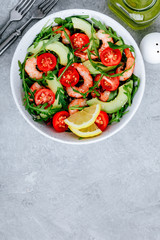 Healthy arugula salad bowl with shrimp, avocado, tomato, and sesame seeds on gray stone background.