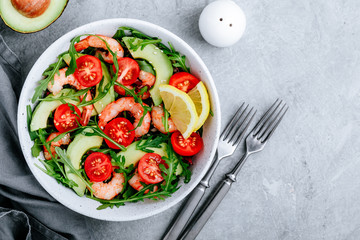 Healthy arugula salad bowl with shrimp, avocado, tomato, and sesame seeds on gray stone background.