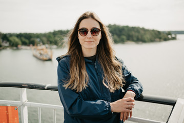 Obraz premium A gorgeous young woman in a blue raincoat and sunglasses poses on the ship in the sunny afternoon 